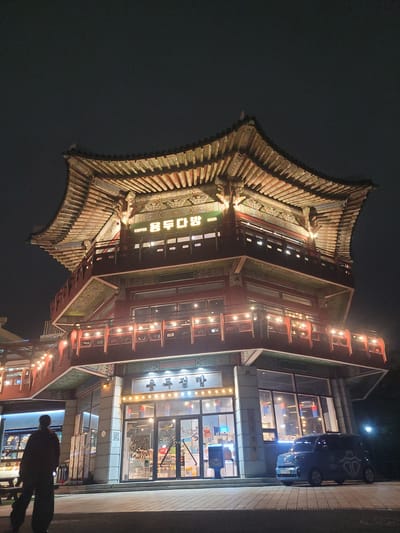 The image depicts a traditional Korean-style building illuminated at night, located in the Haeundae District of Busan, South Korea. The building features multiple tiers with curved, tiled roofs and ornate decorations. String lights are draped along the balconies and the eaves of the structure, creating a warm ambiance.

At the entrance level, large glass windows reveal the interior of what appears to be a cafe or restaurant, with tables and chairs visible. A blue van is parked in front of the building. To the left, a person is walking away from the camera, silhouetted against the lit interior of an adjacent establishment. The ground is paved with interlocking bricks. The sky is dark, indicating nighttime.