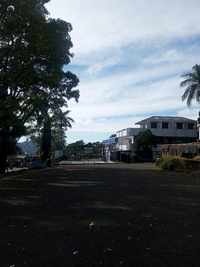 The image captures an outdoor scene in Phonengam, Laos, under a sky with scattered white clouds on a light blue background. The foreground consists of a dark, unpaved or asphalted surface, heavily shadowed, with several small, light-colored pieces of debris scattered across it. To the left, a large, mature tree with dense green foliage fills the frame vertically. Below and behind this tree, a paved road is visible, featuring several parked motorbikes and faint outlines of human figures near what appears to be small market stalls or structures. Further back on the left, hills are discernible on the horizon. In the mid-ground, a metal-fenced enclosure, possibly for a sports court, is present. To the right, a multi-story building complex with white and light gray facades is prominent, featuring multiple dark-paned windows and sections with blue accents. A tall palm tree stands partially visible on the far right. The lower right side of the frame shows a raised embankment with dried vegetation and some wooden elements. No specific actions or interactions are clearly visible among the distant figures.