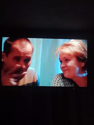 Two individuals, a man and a woman, are visible in this television screen capture. The man, on the left, has a bald head and is looking down and to the left, with his hand near his mouth. The woman, on the right, has short, blonde hair and is looking to her left and smiling. She is wearing a patterned top. The background appears to be a wall with blue and white striped curtains. The image is a screenshot from a television. The location of Chemuzhivka, Ukraine is not evident in this image.