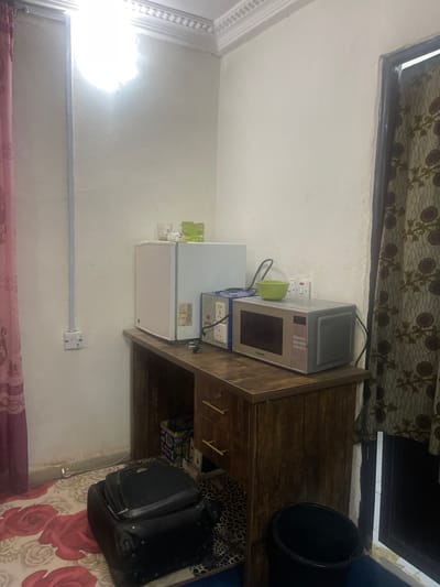 In Kporva, Nigeria, a small room features a wooden desk against a textured cream-colored wall. On the desk, a white mini-refrigerator is next to a gray and silver microwave. A green bowl rests on top of the microwave. Various electrical outlets and a power adapter are visible on the wall and desk. To the left, a pink curtain hangs, and a black suitcase sits on a floral rug. A black trash can is positioned to the right of the desk. A patterned curtain adorns a doorway on the far right. The room appears utilitarian and compact.