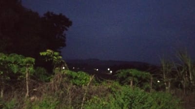 This media file depicts a nighttime landscape scene in Buketteukuh, Indonesia. The foreground is dominated by lush green vegetation, including trees and dense bushes, with some dry, tangled branches in between. Several thin, vertical wooden posts are also visible, suggesting a fence or some form of man-made structure within the natural environment. In the mid-ground, a line of distant lights are scattered across a darkened, hilly terrain, indicating a settlement or populated area. The sky above is a deep, dark blue, characteristic of nighttime. The overall impression is one of a rural or semi-rural area at dusk or after dark, with a sense of quietude. There is no discernible activity or event taking place. The image quality is somewhat grainy, common for low-light photography. No text is visible in the media file.