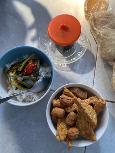 In Madiun, Indonesia, a meal is laid out on a tiled surface, bathed in dappled sunlight casting distinct shadows. The arrangement features three bowls. The first, a blue bowl, contains white rice topped with stir-fried vegetables, possibly water spinach and green beans, seasoned with a vibrant red chili paste. A metal spoon rests in this bowl. Adjacent to it, a white bowl is filled with fried tempeh pieces and what appear to be small, fried meatballs or nuggets. To the right, a clear plastic bag holds several pieces of pale, puffed crackers. In the background, a small glass with a dark liquid, likely coffee, is covered by an orange lid and placed on a glass saucer. The overall scene depicts a casual, home-style meal.