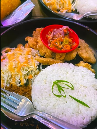 A close-up, eye-level shot presents a meal served on a dark plate, likely in Cadek, Indonesia. The dish features a generous portion of white rice, garnished with a green scallion curl. To the left of the rice is a pile of shredded cabbage and carrots, drizzled with an orange sauce. Beneath this salad, a scattering of crispy brown crumbs is visible. Behind the salad, several pieces of fried chicken are arranged. A small red bowl, positioned above the chicken, contains a spicy relish made of chopped chilies and onions. The meal is accompanied by a fork and spoon, resting on the side of the plate. In the background, a hint of another dish, possibly breaded chicken, and a white paper wrapper can be seen. The lighting appears to be ambient, possibly from indoor restaurant lighting, casting a soft glow on the food.