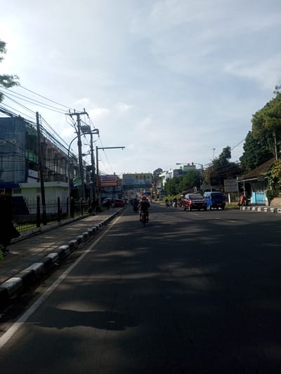 The image depicts a street scene in a town. There is a road, with a motorcycle and some cars in the distance. There are buildings and sidewalks on the sides of the road. Power lines crisscross overhead. A billboard is visible in the background. People are walking on the sidewalk.
