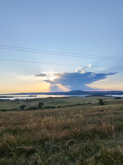 A wide-angle, low-angle shot captures a golden hour landscape scene, likely taken from a hilltop overlooking Tvarditsa, Bulgaria. The sky is a clear, soft blue, transitioning to a warm orange glow near the horizon, where the sun is partially obscured by dramatic, dark clouds. Sunbeams pierce through the clouds, casting ethereal rays across the sky and illuminating the water below.

The foreground is dominated by a field of dry, golden-brown grass, typical of late summer or early autumn. The texture of the grass is detailed, with individual stalks and seed heads visible, some catching the sunlight. In the mid-ground, rolling hills are covered with a patchwork of green and golden fields, suggesting agricultural activity. A winding dirt path is visible on one of the hills, leading towards the water.

A large body of water, possibly a reservoir or a lake, stretches across the middle ground, reflecting the colors of the sky. Across the water, the distant shoreline is lined with rolling hills and a cluster of buildings, indicative of a town or city. Two thin, dark power lines stretch horizontally across the upper portion of the frame, adding a man-made element to the natural scenery. No people or animals are visible in the image. The overall impression is one of serene natural beauty at the end of the day.