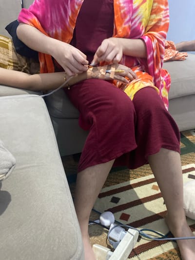 In Walennae, Indonesia, a person's arm is visible, with an IV line attached. The arm is resting on a light-colored sofa, and the IV tubing extends towards the foreground. The person is wearing a maroon-colored garment. Another person, partially visible, is wearing a brightly colored tie-dye shawl in shades of pink, orange, and yellow, also appearing to be seated. The background shows a patterned rug and other furniture.