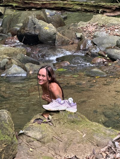 A woman is positioned on a rock outcrop next to a stream, possibly in Suối Giàng, Vietnam. She is smiling and looking directly at the camera. She has long brown hair styled with braids. Next to her are a pair of lavender-colored athletic shoes. The stream flows over rocks and through a lush, green environment. The scene is natural and appears to be outdoors.