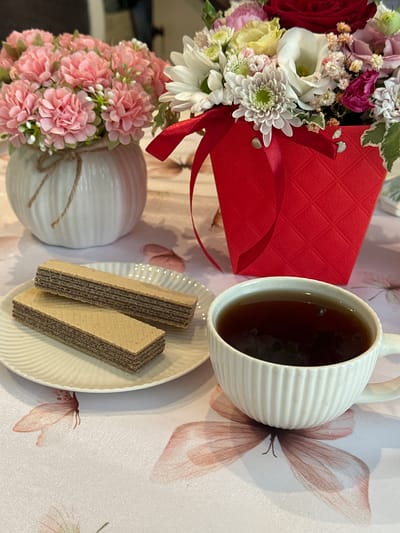 A cup of dark tea sits on a table next to a plate of wafers. A bouquet of flowers in a red box and another bouquet in a white vase are also on the table. The table is covered with a cloth that has a butterfly pattern. The scene appears to be indoors, possibly in a home or cafe setting. There is no indication of the specific location within Wisła, Poland.