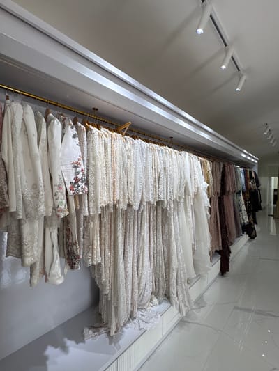 The image shows a retail clothing store, likely a boutique specializing in formal or bridal wear. Racks of elegant dresses, primarily in white and light pastel colors, are displayed along a wall. The garments appear to be made of delicate fabrics like lace and silk, with intricate embellishments such as sequins and embroidery. The store has a clean, modern aesthetic with white walls, a white ceiling, and a polished white tiled floor. Track lighting is installed on the ceiling, illuminating the display. The overall impression is one of sophistication and luxury. The location is identified as Tanjungmeru, Indonesia. There are no people visible in the image.