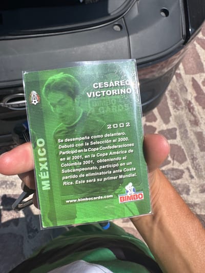 A close-up photograph captures a human right hand holding a green-colored trading card, protected by a clear plastic sleeve, outdoors in the city of León, Mexico. The card prominently features a green-tinted, pixelated portrait of Mexican football player Cesáreo Victorino, with his name and the year "2002" displayed. Text in Spanish details his career as a forward, mentioning his 2000 national team debut, participation in the 2001 Confederations Cup and Copa América, and an upcoming first World Cup. The card includes the Mexican Football Federation logo and the BIMBO brand logo with its website. The individual holding the card wears a green shirt over a white undershirt; a metal carabiner clip is visible attached to a ring near their fingers. The background reveals the rear bumper and partially visible trunk of a dark-colored Nissan vehicle, parked on a light-colored brick or paver surface under strong daylight, which casts shadows.