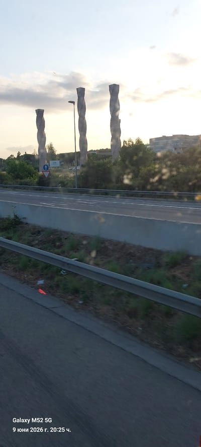 The image shows a view from a vehicle looking towards three large, twisted, sculptural towers in Cerdanyola del Vallès, Spain, on June 9, 2026, at 20:25. The towers, which appear to be made of a light-colored material, are situated in a grassy area with trees and some buildings in the background. A highway or main road runs in the foreground, separated by a concrete barrier and a metal guardrail. The sky is partly cloudy with the sun setting, casting a warm light. Text overlay in the lower left corner indicates the device used, "Galaxy M52 5G," and the date and time. There are no people visible in the image.