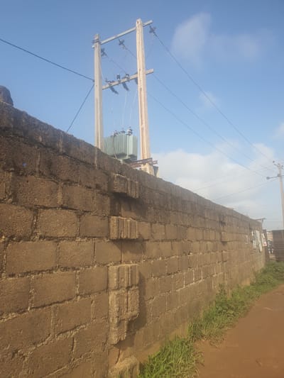 The image depicts an outdoor scene in Unguwar Saya, Nigeria, under a clear blue sky with some scattered clouds. The perspective is tilted, giving a sense of looking upwards and to the side. The primary subjects are an electrical transformer mounted on utility poles, a weathered brick wall, and a paved pathway.
The transformer, a large, light green metal box with cooling fins, is positioned on top of wooden utility poles that support electrical wires. These wires stretch across the frame, disappearing into the distance. The utility poles appear aged, with visible hardware and insulators.
Adjacent to the utility poles and running along the right side of the image is a substantial brick wall. The bricks are rough, irregular, and appear to be made of a dark, earthy material. The wall is partially covered in moss or a similar growth, indicating its age and exposure to the elements. Towards the bottom of the wall, there are some sections where the bricks have deteriorated or are missing, revealing the structure beneath.
In the foreground and extending to the left of the wall is a pathway paved with square-shaped bricks, laid out in a regular pattern. Weeds and grass are growing along the edges of the pathway and at the base of the wall, suggesting that the area might be somewhat unkempt or that nature is reclaiming parts of it.
The scene appears to be set during the daytime, likely late morning or afternoon, given the bright sunlight casting shadows. The overall impression is one of an everyday utility installation in a developing area, with a focus on the infrastructure rather than any human activity. There is no visible text in the image.