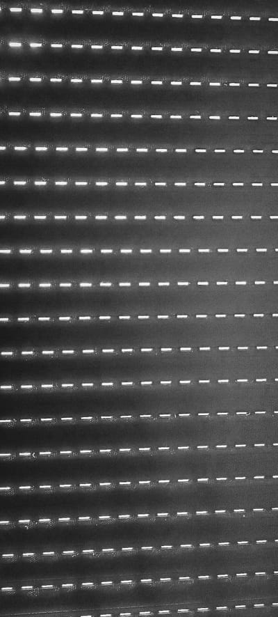 This is a grayscale, close-up, abstract image of a dark, textured surface with a repeating pattern of horizontal white dashes. The dashes appear to be illuminated, suggesting they might be perforations or slots through which light is passing. The texture of the dark background is visible, showing a fine, granular or woven pattern. The dashes are consistently spaced within horizontal rows, and the rows themselves are also evenly spaced, creating a strong visual rhythm and a sense of depth. The image is dark overall, with the bright dashes being the primary source of light. There is no indication of people, specific objects, or a discernible scene. The abstract nature of the image, combined with the repetitive pattern and lighting, creates a minimalist and somewhat austere aesthetic. The context of Winnenden, Germany, does not provide specific visual cues within the image itself to identify the location. The time of day and weather are not discernible. There is no visible text in the image. The image focuses solely on the geometric pattern and texture of the material.