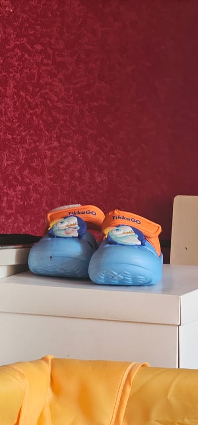 The image shows a pair of blue children's flip-flops with orange straps, each featuring a cartoon character. The flip-flops are placed on a white surface, likely a shelf or table, next to a red textured wall. To the right of the flip-flops, a bright yellow fabric is draped, partially obscuring the white surface. The text "TikkaGO" is visible on the orange straps of the flip-flops. The lighting suggests an indoor setting. There are no people or clear indications of time of day or weather. The location context provided is Kobryn, Belarus.