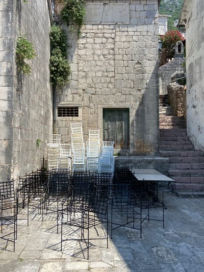 The image depicts a sunlit outdoor courtyard in Perast, Montenegro, adjacent to an old stone building. Piles of white plastic chairs are stacked to the left, in front of a window with a metal grate and a weathered green wooden door. In the foreground, numerous black metal chairs and tables are arranged, casting long shadows on the stone-tiled ground. To the right, a stone staircase ascends, leading to a higher level. The walls are constructed of rough-hewn stone blocks. Green ivy grows on the left wall and a small pink-flowered bush is visible near the top of the stairs on the right. The scene appears to be set up for outdoor dining or an event.