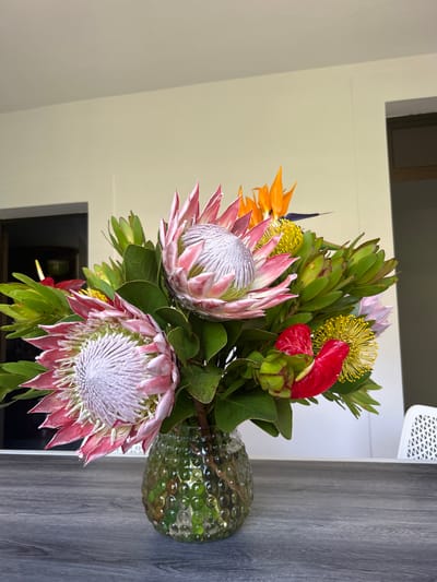 A vertical, medium-shot captures a floral arrangement in a speckled glass vase. The arrangement consists of two protea flowers with pink petals and white centers, accented by yellow pincushion flowers, an orange bird of paradise, and red anthurium flowers. Green foliage surrounds the flowers. The vase sits on a grey, wood-grain surface. The background consists of a light beige wall and a glimpse of a black object. The image does not appear to have been taken in Câmara de Lobos, Portugal; the setting is indoors and does not showcase features of the city.