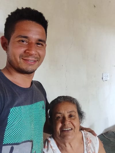 An elderly woman with gray hair and a white floral blouse smiles warmly. Next to her, a younger man with short dark hair and a green-striped dark t-shirt smiles widely, displaying his teeth. The background features a plain white wall with a visible crack and a white electrical outlet.