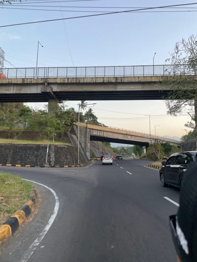 The image depicts a road with two vehicles and a pedestrian overpass in Maumbi, Indonesia. The overpass, constructed of concrete and metal railings, spans over the road. A street light is visible on top of the overpass. The road itself is paved and marked with white lines, and features a grassy embankment on the left side with yellow and black striped barriers. On the road, a black car is seen in the foreground on the right, and a white car is in the distance on the left, along with two other vehicles and a motorcycle. The scene is set against a backdrop of trees and hills, under a pale sky. The overall impression is that of a typical urban or suburban transportation artery.