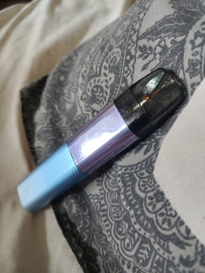 An electronic cigarette is lying on a patterned fabric. The e-cigarette is cylindrical with a black, translucent mouthpiece. The body of the e-cigarette is divided into two sections: the bottom section is light blue with a textured grid pattern, and the top section is iridescent and reflects purple and silver. The fabric beneath the e-cigarette is a light beige color with a repeating grey paisley pattern. The image is a close-up shot, with the e-cigarette positioned diagonally across the frame. There is no indication of the city of Okhtyrka, Ukraine in this image.
