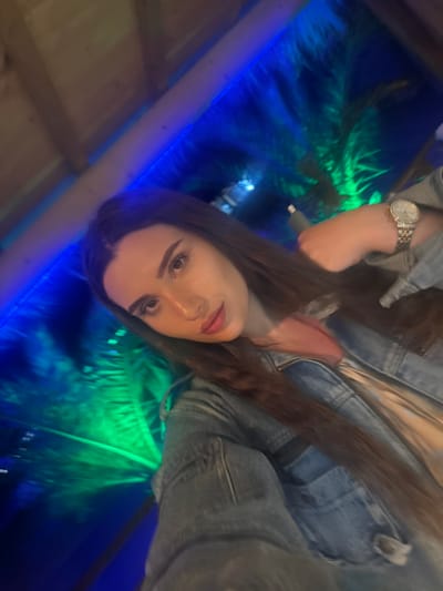 In an indoor setting illuminated by vibrant blue and green lights, a young woman with long, dark brown hair takes a selfie. She is wearing a denim jacket over a pink top, and a silver watch is visible on her left wrist. Her gaze is directed towards the camera, with her head tilted slightly to the side. The background suggests a tropical or jungle-themed environment, possibly a bar or club, with blurred foliage illuminated by colored lights. The ceiling appears to be made of wood. The location is the city of Primorsko, Bulgaria.