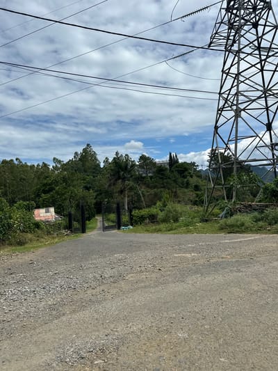 The image shows a rural landscape with a dirt road leading towards a gate. To the right, a large electrical tower dominates the scene. The road is rough and unpaved, with gravel and dirt. Beyond the gate, the road curves into a densely wooded area with many trees and some bushes. In the distance, a hill rises, topped with a few buildings and more trees. The sky is partly cloudy, with patches of blue sky visible between the clouds. The overall impression is one of a remote, undeveloped area.