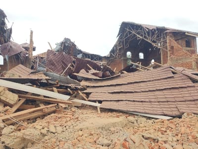 The scene depicts the extensive remnants of a collapsed brick building in Beni, Democratic Republic of the Congo. The foreground is dominated by a deep pile of reddish-brown brick rubble, concrete fragments, and soil. Interspersed within this debris are multiple large, corrugated metal roof panels, predominantly brown, along with numerous fractured wooden beams and planks. In the midground and background, the primary structure, constructed of red brick, shows catastrophic failure. Its entire roof system, composed of wooden trusses and brown metal sheeting, has collapsed inward and outward, exposing the skeletal framework of the upper walls. Distinct arched window openings are visible in the partially standing brick walls. A fractured brick column or chimney-like structure remains upright amidst the wreckage on the left. The sky is uniformly overcast. No individuals or active elements are present.