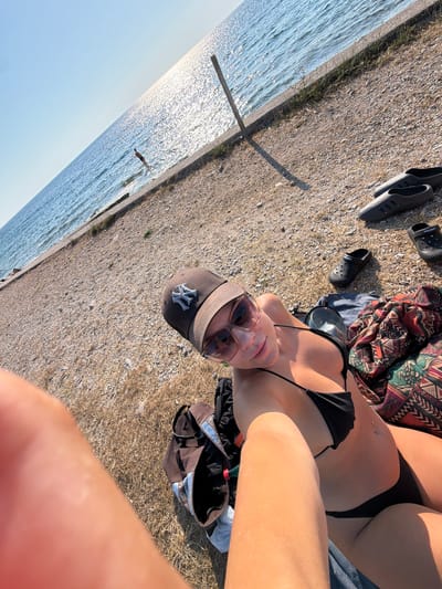 This media file captures a person lounging by the water on a sunny day. The scene appears to be a beach or waterfront area, with a pebbly ground and the glistening surface of the sea. The person is lying on their back, wearing a black bikini and sunglasses, with a baseball cap on their head. A colorful patterned blanket and a black bag are nearby, along with a pair of black Crocs. The sun is casting shadows, indicating it's likely daytime, possibly late afternoon given the angle of the light. The water is a clear blue, and the sky is bright. The overall mood seems relaxed and summery, suggesting a day of leisure by the sea. The baseball cap has a visible "NY" logo.