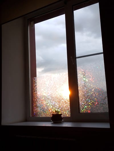 The image depicts a window with a view of an overcast sky and the sun breaking through the clouds. The window is divided into two panes, both of which are covered with holographic film that refracts light into a spectrum of colors. On the windowsill, a small potted plant is visible. The scene is set in an indoor environment, with a dark wall on the left and a dark curtain on the right. The lighting suggests it is either dawn or dusk. This window is located in the city of Birsk, Russia.