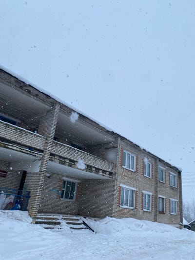 A tilted view looking down on a brick apartment building during a snowfall. The building has several floors with white-framed windows. Balconies extend from each floor, also constructed of brick. Heavy snow accumulation is visible on the ledges, roof, and ground. The sky is overcast and filled with falling snow.