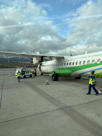 An active airport tarmac scene unfolds during the daytime under a partly cloudy sky, featuring an aircraft and ground personnel. The main subject is a turboprop aircraft, distinctively painted with a white fuselage, a prominent green stripe along its lower half, and a thinner blue stripe above it. The airline's name, "BINTER LÍNEAS AÉREAS DE CANARIAS," is visible along the top of the fuselage, while "bintercanarias.com" is printed below the windows.
Two ground crew members, both wearing high-visibility yellow vests and dark blue pants, are attending to the aircraft. One female crew member is positioned near the front of the plane, intently looking down at a tablet or device she holds in her hands, suggesting a pre-flight or post-flight check. Further towards the rear of the aircraft, another male crew member stands beside a white ground support vehicle, which has "inter" written on its side (likely part of "Binter"). An orange traffic cone is placed near the front landing gear of the plane.
The ground is a grey concrete tarmac, showing some tire marks or wear. In the background, beyond the plane's wing and tail, a sprawling landscape of mountains or hills is visible, dotted with buildings, under a mix of blue sky and scattered white and grey clouds. The overall impression is one of routine airport operations, likely in the Canary Islands, given the airline's name and the suggested location of Granadilla de Abona, Spain.