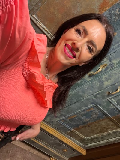 A woman smiles brightly at the camera in an indoor setting in Konz, Germany. The image, oriented vertically, captures her from the chest up. She has dark, shoulder-length hair and is wearing a vibrant coral or peach-colored top with a smocked bodice and ruffled neckline. A silver-toned chain necklace, featuring a distinctive interlocking link, adorns her neck. Her left hand, with light-colored nail polish, is partially visible in the upper left.
Her expression is joyful and engaging, with a wide smile revealing bright pink lipstick and white teeth; a small, shiny detail is visible on one of her upper teeth. Her eyes are dark and enhanced with makeup.
The background consists of what appears to be an antique-style piece of furniture or paneling, possibly a cabinet or door. It features a distressed blue-gray finish with gold accents, including raised panels and decorative handles. To her left, a warm-toned wall or wooden surface with additional golden trim is visible. The lighting suggests an indoor environment. No text is visible in the frame.