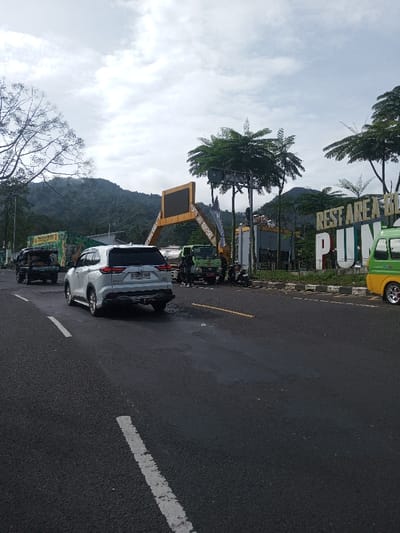 The image captures a daytime scene on an asphalt road with a mix of vehicles, set against a backdrop of lush green mountains under an overcast sky.
In the foreground, a dark gray or black asphalt road stretches from the bottom towards the middle of the frame. A broken white line is visible on the left side of the road, indicating a lane divider. The road surface appears somewhat wet or reflective in patches, possibly from recent rain or oil spills, particularly under the white SUV.
Towards the left side of the mid-ground, a white SUV with an Indonesian license plate "B 1973 QWH" is prominent, facing away from the viewer. It appears to be moving or recently stopped. To its left, partially obscured, is the back of what looks like a dark green or black pickup truck with an open bed, possibly carrying goods.
On the right side of the road, beyond a low curb, there's an area with more vehicles and structures. A large sign with the text "REST AREA B..." and "PUN..." (the latter likely part of "Puncak," a common rest area name in Indonesia) is visible. The letters are bold and appear to be painted in shades of green, yellow, and red, or are illuminated, standing out against a dark background. To the right of this sign, a yellow and green minivan is parked or moving slowly, with custom chrome rims on its wheels.
Further back, near the "REST AREA" sign, a green truck with a tanker or similar body is parked. Several individuals appear to be gathered around this truck and a couple of motorcycles parked nearby, suggesting a stop or a small gathering. Behind them, a large rectangular digital billboard with a dark screen is mounted on a distinctive yellow or gold-colored arch-like structure.
The background is dominated by a range of green, forested mountains, indicating a hilly or mountainous region. The sky above is overcast with soft, diffuse light, suggesting a cloudy day. Bare branches of a large tree are visible on the far left of the image, extending upwards into the sky. Scattered trees with green foliage are also visible throughout the scene, particularly to the right of the billboard and around the rest area sign.