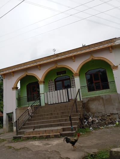 The image shows a two-story building with a concrete staircase leading up to a set of double doors. The building is painted white with a yellow trim, and has a green painted wall below the windows. The building has a series of arches in the facade, with some of the windows open. The building has a large, gray concrete wall under it. There are power lines running overhead. A brown and white rooster is walking at the bottom of the stairs. The building appears to be a mosque.  The location is at the coordinates (latitude: -6.971, longitude: 107.519) in Kecamatan Kutawaringin, Indonesia.