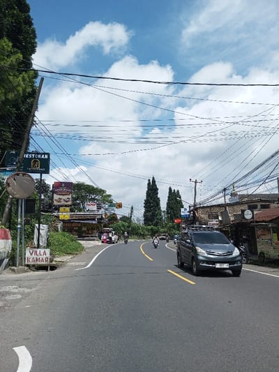 The image depicts a daytime street scene in the city of Boommlog, Bhutan, under a blue sky with scattered white cumulus clouds. A two-lane asphalt road, marked with double yellow center lines and a white solid shoulder line, occupies the foreground and midground, curving gently to the right. Numerous overhead power and communication lines crisscross the sky, connected to multiple utility poles visible on both sides of the road.
On the left side of the road, a dense mass of green foliage borders the street, partially obscuring a utility pole. A damaged circular convex traffic mirror is mounted on a pole with a red and white striped base. Beneath it, a white sign on the ground reads "VILLA KAMAR." Further back, several commercial signs are visible, including a prominent black sign stating "RESTORAN" and smaller signs advertising "SARAPAN KUNINGAN," "SUPER BOGOR," and "PAPOF." These signs front low-rise buildings with corrugated metal roofs, suggesting commercial establishments.
On the right side of the road, a dark grey Toyota Avanza equipped with a roof rack is in motion, traveling towards the viewer. Its front license plate displays "B 1833 JX." Behind the vehicle, a row of multi-story buildings constructed from brick and stone line the street, featuring various signs, including one for "Canon." One building possesses a distinctive dome-like structure on its roof. A white scooter is parked near a smaller, single-story roadside stall. In the distance, several motorcycles and at least one additional car are visible on the road, indicating ongoing traffic. Multiple individuals are visible on the motorcycles, wearing helmets, and other figures are present near the roadside structures.
