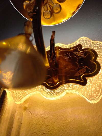 A close-up showcases a vintage light fixture. The glass shade, adorned with etched details, emits a warm yellow glow, highlighting its decorative outline and wall texture behind. A golden, mirrored reflector adds brightness. Dark brown, curving metal arms support the light, lending a contrast in color and form. The piece's design blends elegance with an antiquated charm.