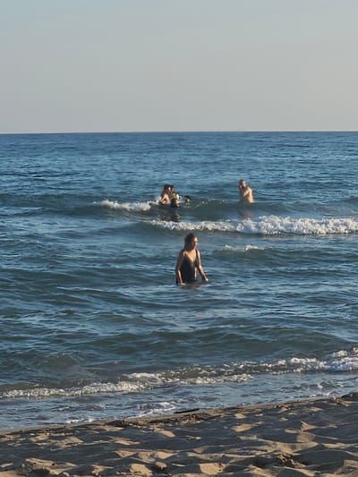 The image shows a group of people in the ocean at Castelldefels, Spain. The orientation of the image is vertical, with the top of the frame showing the sky and the bottom showing the sand. The water is a clear blue, with visible waves and foam. The sand appears to be light brown and textured, with distinct patterns from what looks like small waves or ripples.

Three individuals are clearly visible in the water. One person is lying on their back, seemingly enjoying the water. Another person, wearing a dark swimsuit, is in a seated position, looking towards the left of the frame. A third person is partially submerged, with their head and arms visible. They appear to be swimming or playing in the water. The presence of people enjoying the ocean suggests a recreational activity, likely on a warm and sunny day, typical for a beach setting in Spain. The sky is a light, pale blue, indicating fair weather and possibly late morning or early afternoon given the shadows cast on the sand. No text is visible in the image.