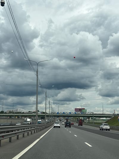 The image shows a highway with multiple lanes, a metal guardrail, and streetlights. Several vehicles, including cars and trucks, are visible on the road, suggesting traffic. In the background, there are buildings and trees, indicating an urban or suburban setting. The sky is filled with dark, heavy clouds, implying overcast or stormy weather. Two red balloons and one white balloon are suspended in the air, attached to wires that run across the frame. The image is oriented vertically, with the highway appearing to extend upwards into the sky. Visible text includes "СТРОЙ ДВОР" and "ВЫХОД", which are Russian words related to construction and exit. The scene appears to be captured from a moving vehicle on the highway.