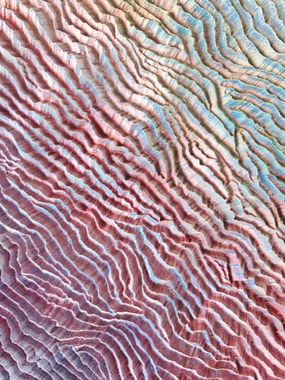 This is a close-up, abstract aerial photograph showcasing a textured landscape. The dominant features are parallel, undulating ridges that create a sense of depth and rhythm. These ridges are colored in shades of pink, red, and blue, with hints of pale green and yellow. The lighting suggests a bright, possibly sunny day, casting subtle shadows that accentuate the contours of the land. The surface appears granular, possibly sand or soil, with a weathered texture. The abstract nature of the image, with its swirling patterns and vibrant, artificial-looking colors, evokes a sense of organic artistry or a stylized, otherworldly terrain. There are no people, objects, or visible text. The context provided places this landscape within the Zhdanovichsky Selsoviet in Belarus, suggesting it is a natural or possibly man-made feature within that region.