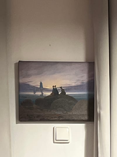 A rectangular canvas print is mounted on a light-colored wall in Capital City of Prague, Czechia. The print depicts a landscape featuring three dark-clad figures seated on large, rounded rocks in the foreground, observing a body of water under a twilight sky. A low light source, appearing as a sun or moon, casts an orange-yellow glow on the horizon. Dark, indistinct landmasses or rock formations rise from the water in the mid-ground. The sky above is characterized by shades of deep blue, purple, and orange. Below the canvas, a white, square light switch with a single push-button is affixed to the wall. A light-colored curtain hangs vertically along the right side of the image, partially obscuring the wall and the right edge of the print. A small dark speck is visible on the wall above the painting.
