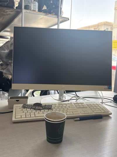 The media file shows a desktop computer, a keyboard, and a cup on a desk. The computer has a cream-colored bezel and a dark screen. The keyboard is also cream-colored, with some of the keys displaying Arabic characters. A blue pen rests on the keyboard, and a watch is placed on the keys. To the right of the keyboard, a dark green cup with a patterned rim is visible. The scene appears to be an indoor setting, possibly an office or study, with a window in the background showing a building and what looks like a street. Sunlight streams in from the window, suggesting it is daytime. The overall impression is one of a workspace. There are some labels on the computer bezel, one appears to say "Intel Core i7" and another "Intel Iris Xe".