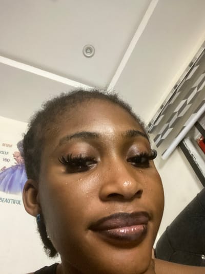 A close-up, low-angle shot shows the face of a young Black woman indoors, with part of a ceiling visible. The woman has dark skin, and her eyes are adorned with long, dramatic false eyelashes. She wears dark lipstick with a glossy finish. To her left, a poster with text is partially visible, with the words "BELIEVE," "YSELF," and "YOU" discernible. Further to the left, a portion of a figure wearing a light purple dress is also visible on the poster. To the right of the woman, a set of blinds with a geometric pattern is present. The woman is wearing a dark top. The lighting in the room appears to be artificial.