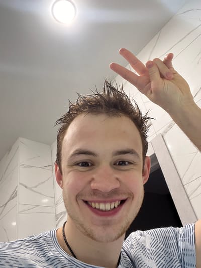 A young adult male is depicted in a close-up selfie taken within a white-tiled interior, consistent with a bathroom environment. His face, angled slightly upwards, occupies the lower half of the frame. He has light brown, wet, spiky hair and a light amount of stubble on his chin and jawline. He is smiling broadly, revealing his teeth, and looking directly at the camera. He is wearing a light-colored t-shirt with thin horizontal stripes and a dark chain necklace. His right hand, extended above his head and to the right, forms a "V" or peace sign with the index and middle fingers raised. The background features white, large-format tiles with subtle grey veining, visible on both a straight wall and a corner to the left. A bright, circular overhead light fixture is prominently visible in the upper center of the frame, casting strong illumination. A dark opening, possibly a doorway or shower entrance, is partially visible on the right. The specific city of Tliustenkhabl', Russia, cannot be determined from the visual information presented in the image.