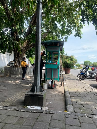 A street scene shows a food cart near a leafy tree. A vendor wearing a white cap stands near the cart. Another person in a yellow shirt is seated on a concrete bench using a phone. A utility pole stands beside the bench. The sidewalk is paved with stone blocks. Motorbikes are parked along the curb in the distance. The sky is overcast.