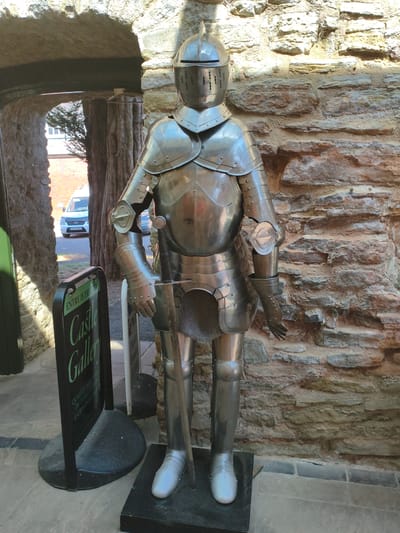 A full-body, life-sized suit of polished medieval armor stands upright on a black pedestal against a rough-hewn stone wall. The armor is detailed, featuring a helmet with a visor, segmented plates on the chest, arms, and legs, and a chainmail skirt. A sword is sheathed in its scabbard, visible between the legs of the armor. To the left of the armor, a black A-frame sign with green text reading "ENTRY IS FREE TO Castle Gallery" is positioned on the cobblestone ground. In the background, an arched stone entryway reveals a glimpse of a white van parked on a street and part of a brick building. The lighting suggests it is daytime, with shadows cast by the sunlight. The scene appears to be in Ludlow, United Kingdom.
