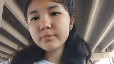 The media file is a close-up selfie of a young woman, likely of East Asian descent, with dark hair and brown eyes. She is wearing a white t-shirt and a small, rectangular earring in her right ear. Her facial expression is neutral. The background consists of a series of parallel, light-colored beams, suggesting an outdoor or semi-outdoor setting, possibly under a bridge or a structure with a similar design. The lighting indicates it is daytime. The overall impression is a casual, unposed snapshot. There is no visible text in the media file.