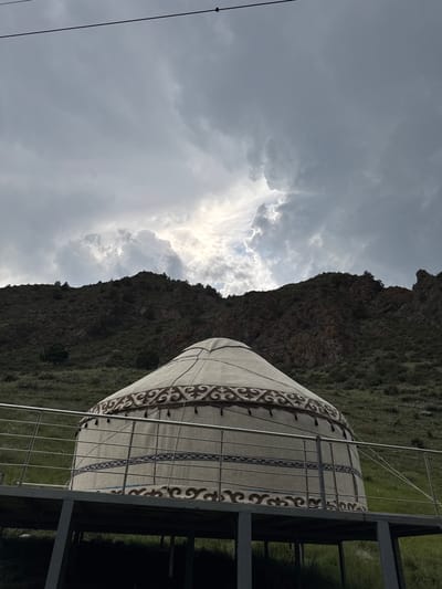 This is a medium shot of a yurt situated on a raised platform with a metal railing in front of a steep, rocky hillside. The yurt is decorated with intricate brown and beige patterns around its circumference and a decorative band around its upper section. The hillside behind the yurt is covered in sparse green vegetation and dotted with trees. The sky above is filled with dramatic, heavy clouds, with a patch of bright sunlight breaking through near the center. A thin power line stretches horizontally across the top of the frame. The scene appears to be in a natural, possibly mountainous, outdoor environment, consistent with the location of Kyzyl-Korgon, Kyrgyzstan. There are no people or animals visible in the image.
