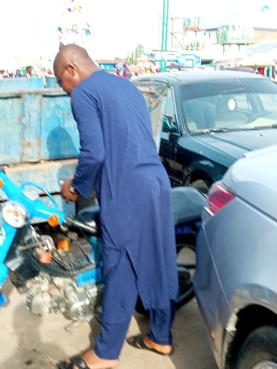 The image depicts an outdoor scene in Arawa, Nigeria, likely in a marketplace or street area. In the foreground, a man wearing a blue traditional garment and sandals is interacting with a bright blue motorbike. The man's hands are near the motorbike's engine, suggesting he might be working on it or inspecting it. The motorbike appears to have parts exposed, indicating it could be undergoing maintenance or is in a state of disrepair. Behind the motorbike and the man, a dark green car is parked closely, with its front windshield and part of its roof visible. Further in the background, a light blue vehicle is positioned to the right of the green car. The environment appears to be an urban setting with stalls or shops visible in the distance, some with colorful awnings, and what looks like a green structure, possibly a pole or part of a building, rising high into the sky. The ground surface is concrete or asphalt. The lighting suggests it is daytime, with the sky appearing bright.