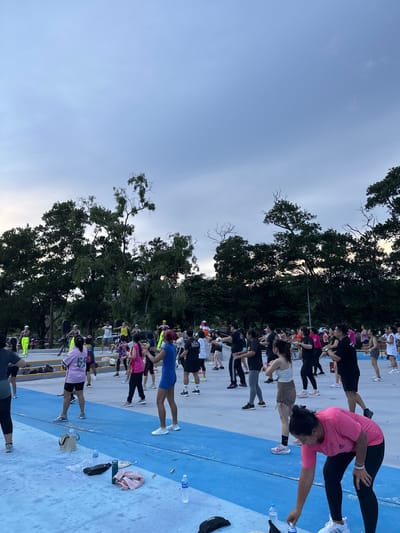 The image depicts a group of people engaged in an outdoor fitness class, possibly a workout or exercise session, at what appears to be a public park or recreational area in Phuket City Municipality, Thailand. The setting features a blue and white tiled ground, bordered by a yellow line, with lush green trees providing a backdrop. The sky is overcast, suggesting either early morning or late afternoon, with the low light casting soft shadows.

The individuals, predominantly women, are dressed in athletic wear in various colors, including pink, black, white, yellow, and blue. They are positioned in a line, performing exercises that involve stretching and core strengthening, such as planks and lunges. Some are holding water bottles, indicating a sustained activity. The overall mood seems focused and energetic, with participants concentrated on their movements. The presence of trees, the specific tiling of the ground, and the likely warm climate implied by the attire suggest a tropical or subtropical location, consistent with Phuket. No visible text is present in the image.