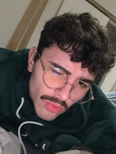 A person with dark, curly hair and a neatly trimmed mustache is wearing round, gold-rimmed glasses. They are posed at a slight angle, looking up. The person is wearing a dark green hoodie with white drawstrings. The background is slightly out of focus, revealing hints of a wooden surface and light-colored curtains. The overall tone is casual and relaxed.