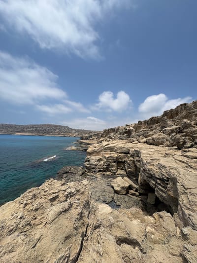 This is a vertical, eye-level shot of a rugged, rocky coastline meeting a clear blue sea under a bright, cloudy sky. The foreground is dominated by a textured, beige rock face, showing deep crevices and layered sedimentary rock formations, suggesting a natural, weathered structure. A dark, shadowy opening within the rock face, resembling a small cave or alcove, is visible on the right.

Beyond the rocks, the turquoise water stretches out, appearing calm with gentle ripples. A narrow strip of darker, rocky land or hills can be seen in the distance along the coastline, leading towards the horizon. The sky is a vibrant blue, dotted with fluffy white clouds, indicating fair weather. The lighting suggests it is daytime, likely late morning or early afternoon, with the sun casting shadows that highlight the rock's texture.

The scene evokes a sense of natural beauty and tranquility, typical of a Mediterranean coastal landscape. There are no people or man-made objects visible, focusing the attention entirely on the geological formations and the sea. The location context places this scene in Ayia Napa, Cyprus, known for its picturesque coastlines. No text is visible in the image.